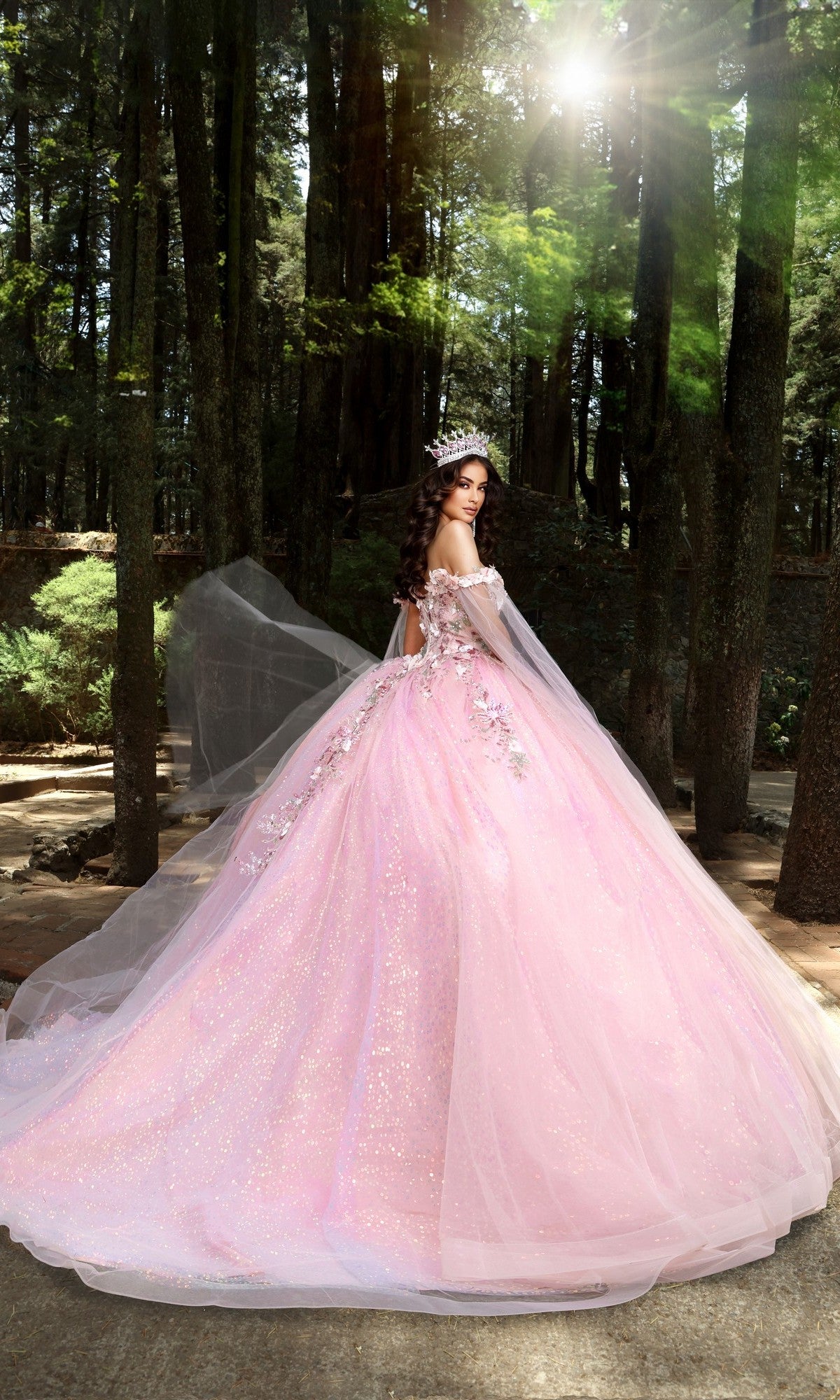 A woman wearing the Quinceanera Dress Q114 by Nina Canacci and a tiara stands in a sunlit forest, looking over her shoulder.